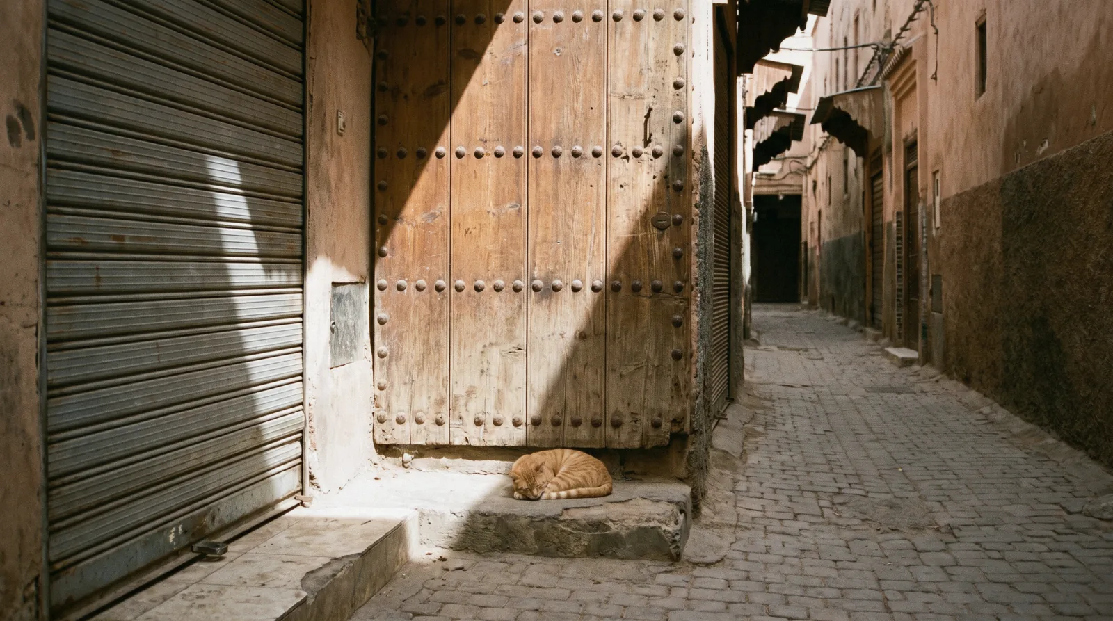 A closed studded cedar door and a shuttered shop in an empty medina lane at midday, the Marrakech opening hours question in one frame